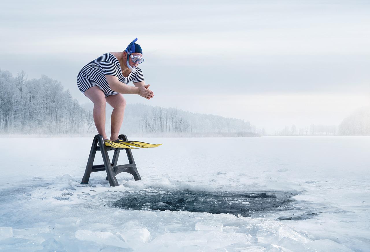 Bereit zum Sprung ins kalte Nass: Eine Person mit Tauchausrüstung und Flossen steht auf einer Leiter und schaut auf das Eisloch im gefrorenen See.